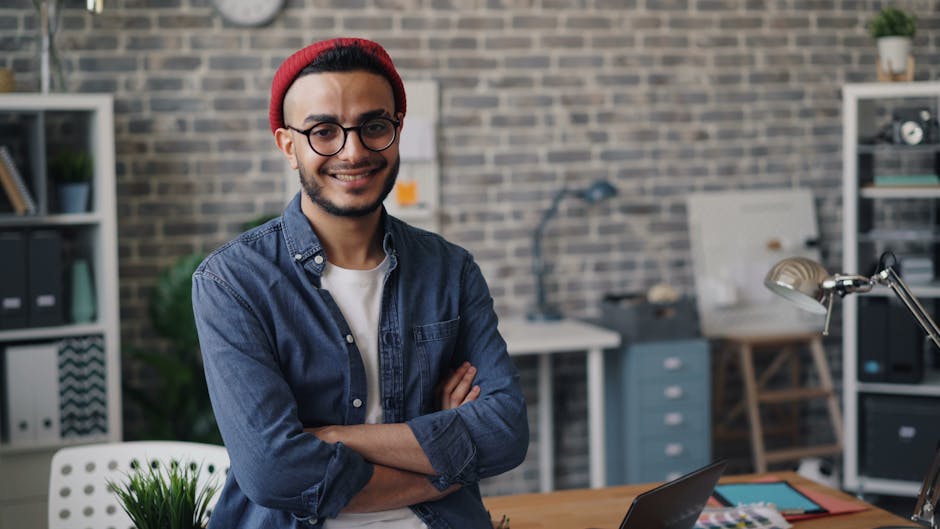 Smiling young man standing confidently in a modern office with a brick wall background.