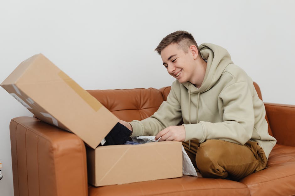 Smiling young man unboxing a package while sitting on a brown leather couch indoors.