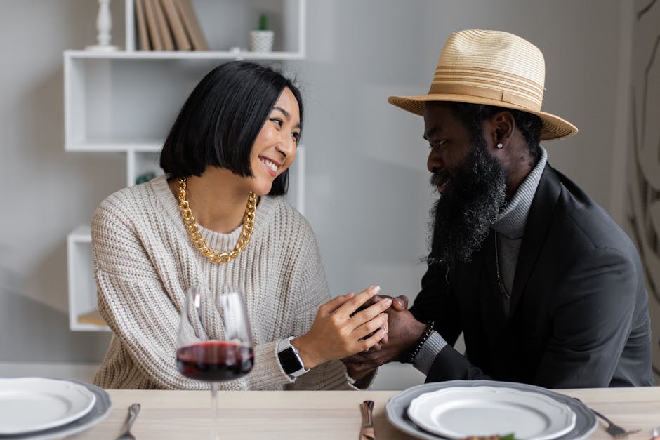 Loving multiracial couple looking at each other holding hands while sitting at table with plates and wine and having dinner