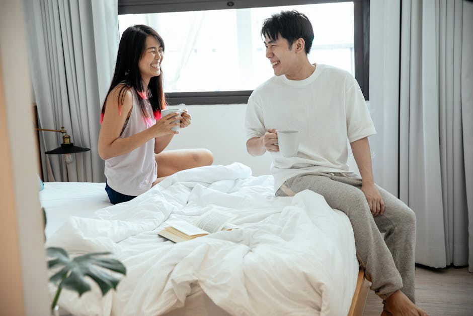Laughing Asian couple looking at each other and enjoying hot coffee in morning while sitting on bed
