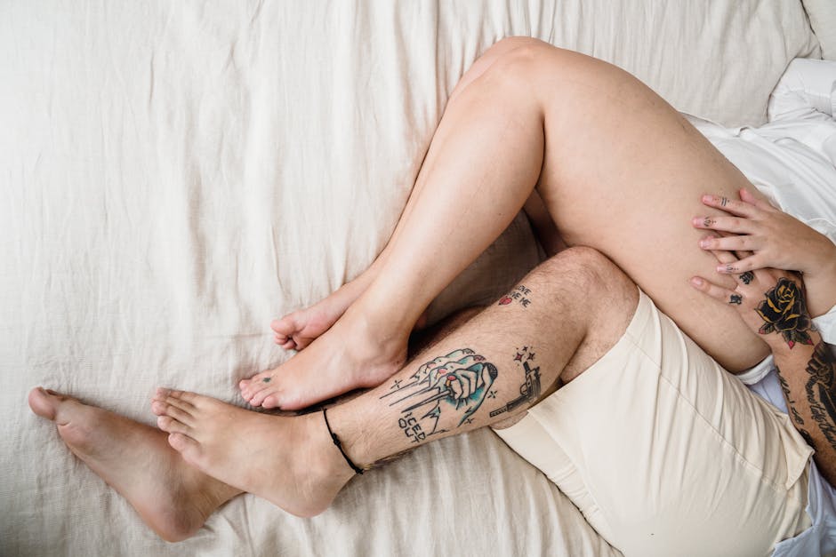 Close-up of a couple's tattooed legs intertwined on the bed in daylight.