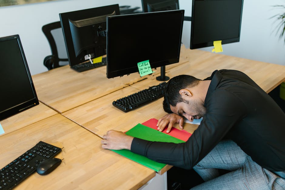 Exhausted office worker falling asleep at desk with monitors and keyboard.