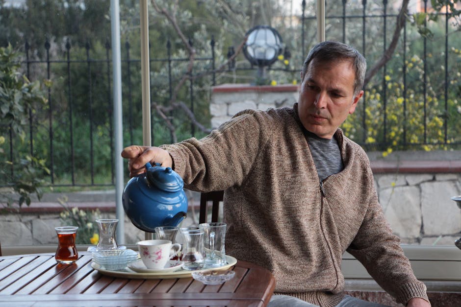 A man pours tea from a blue teapot on a patio table surrounded by glassware.