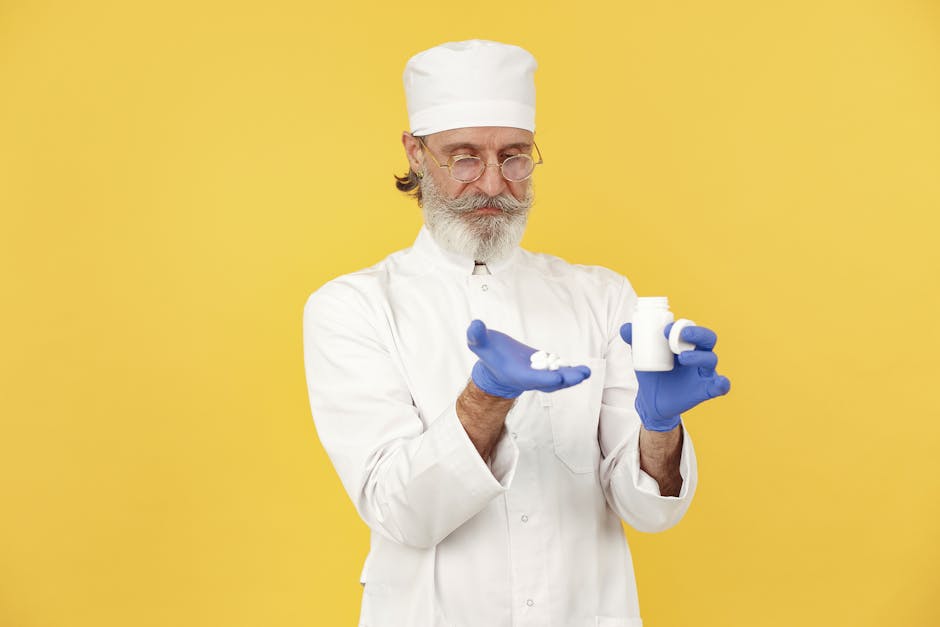 Senior pharmacist with beard and glasses holding medicine bottle and tablets on a yellow background.