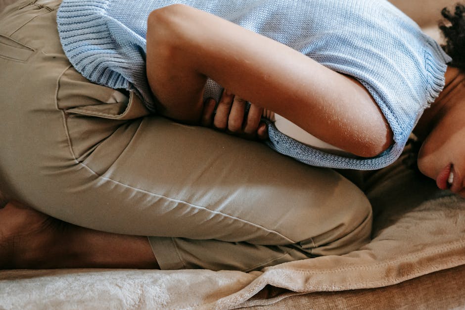 A young woman in discomfort, holding her stomach while curled up on a couch indoors. 